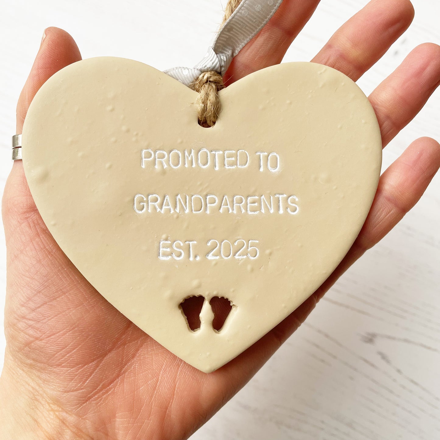 Beige clay hanging heart with baby feet cut out of the bottom and white personalisation, the heart is personalised with PROMOTED TO GRANDPARENTS EST. 2025