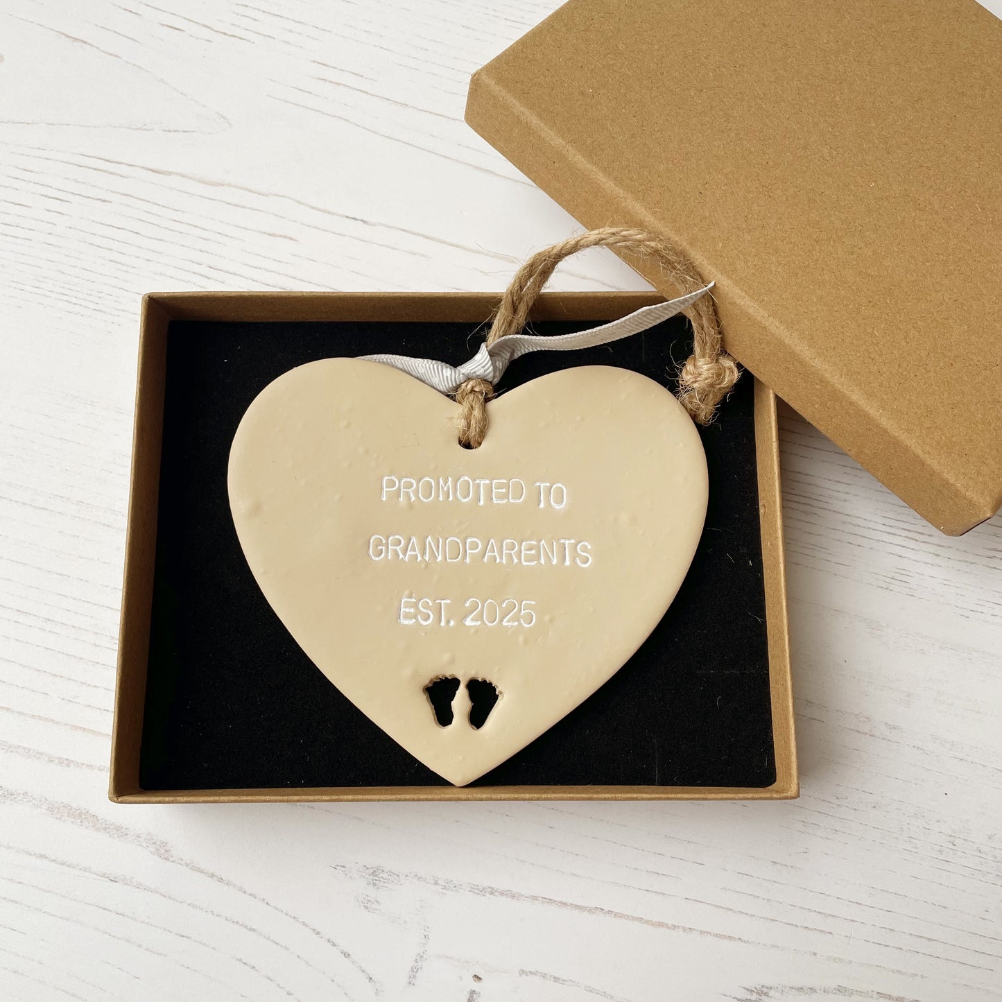 Beige clay hanging heart with baby feet cut out of the bottom and white personalisation, the heart is personalised with PROMOTED TO GRANDPARENTS EST. 2025