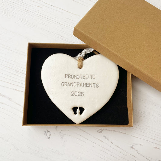 Pearlised white clay hanging heart with baby feet cut out of the bottom and grey personalisation, the heart is personalised with PROMOTED TO GRANDPARENTS 2025