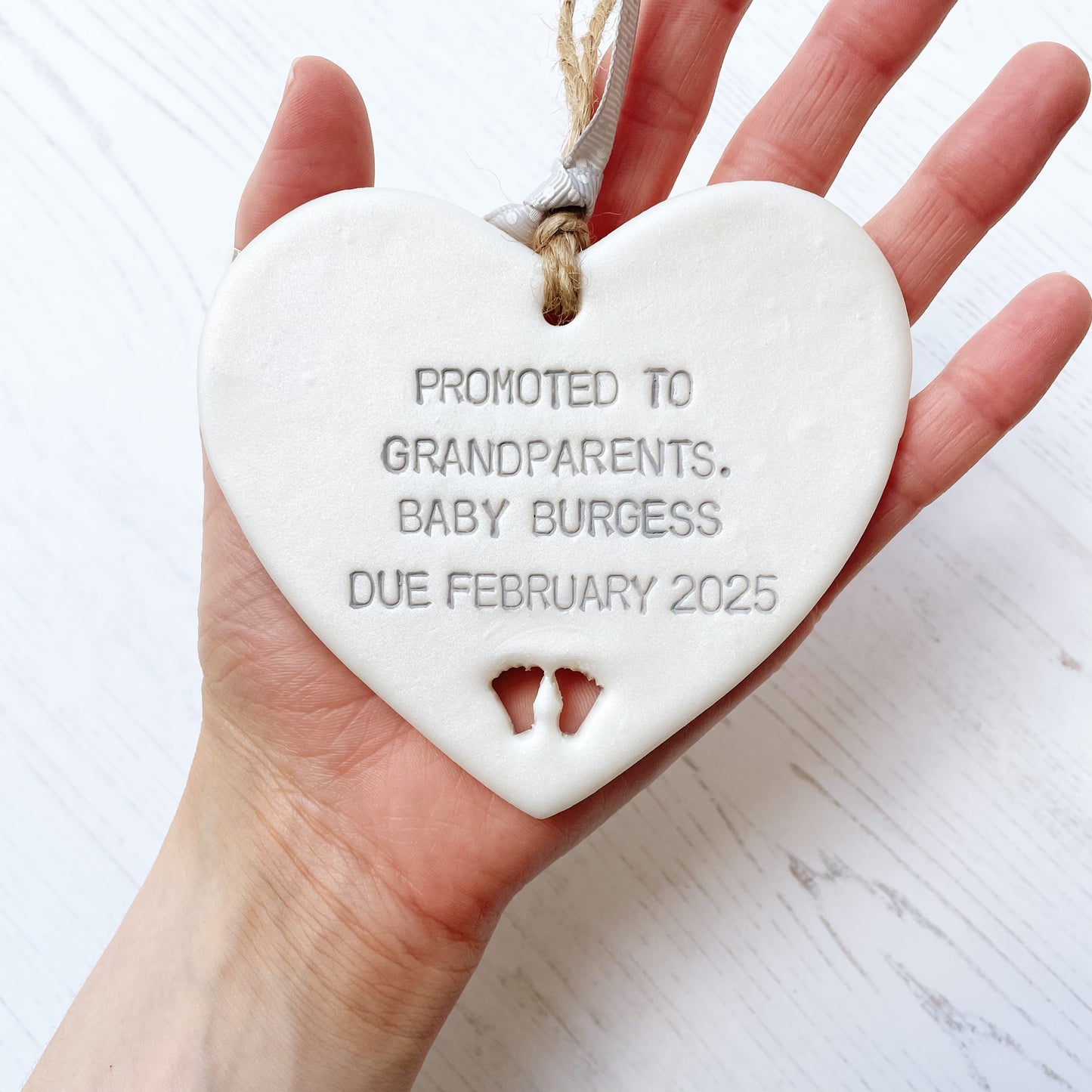 Pearlised white clay hanging heart with baby feet cut out of the bottom and grey personalisation, the heart is personalised with PROMOTED TO GRANDPARENTS. BABY BURGESS DUE FEBRUARY 2025