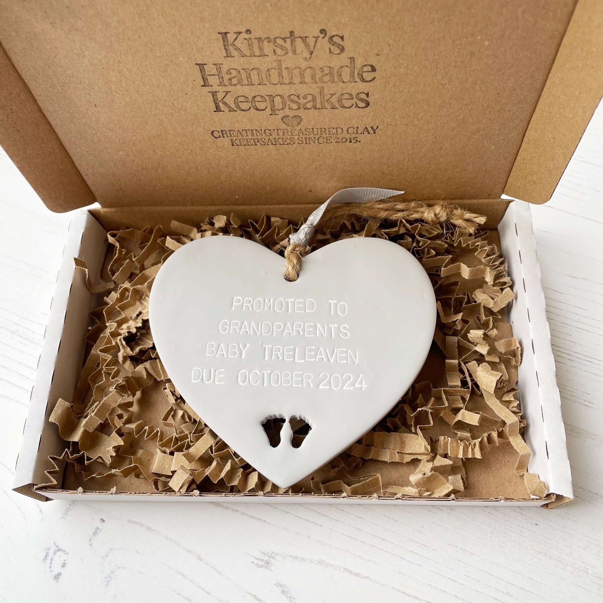 Grey clay hanging heart with baby feet cut out of the bottom and grey personalisation, the heart is personalised with PROMOTED TO GRANDPARENTS BABY TRELEAVEN DUE OCTOBER 2024