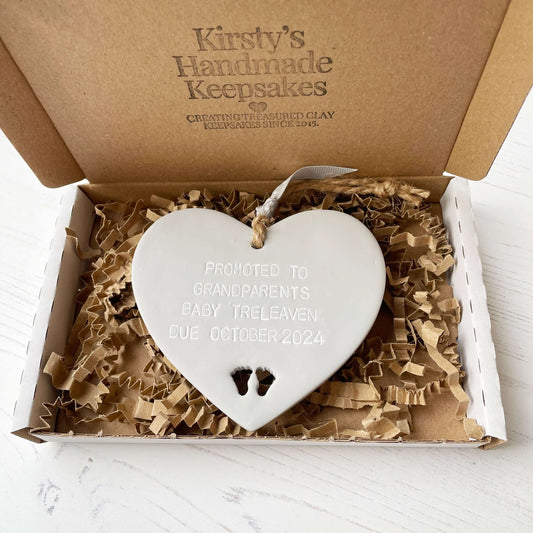 Grey clay hanging heart with baby feet cut out of the bottom and grey personalisation, the heart is personalised with PROMOTED TO GRANDPARENTS BABY TRELEAVEN DUE OCTOBER 2024
