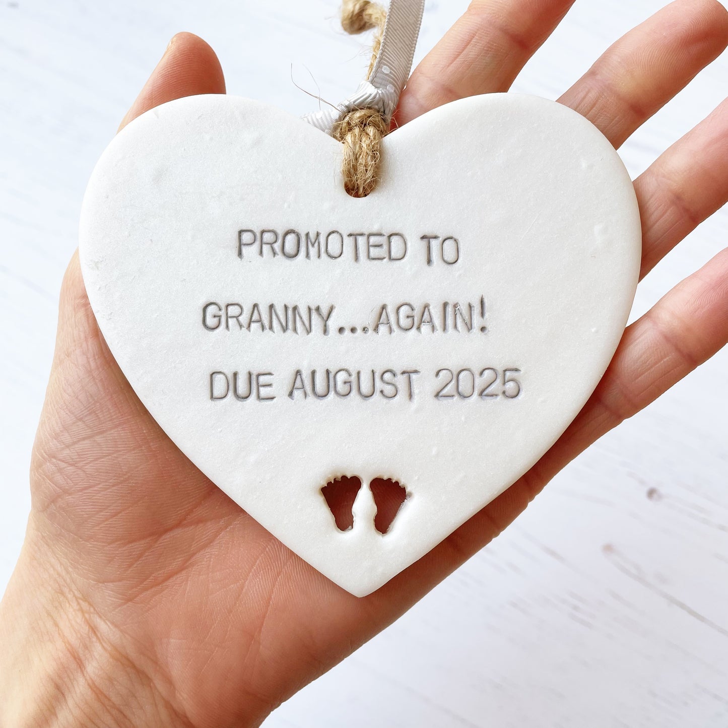 Pearlised white clay hanging heart with baby feet cut out of the bottom and grey personalisation, the heart is personalised with PROMOTED TO GRANNY… AGAIN! DUE AUGUST 2025