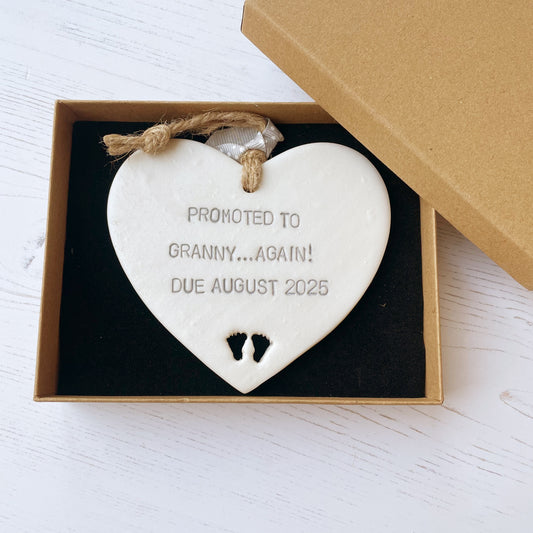 Pearlised white clay hanging heart with baby feet cut out of the bottom and grey personalisation, the heart is personalised with PROMOTED TO GRANNY… AGAIN! DUE AUGUST 2025