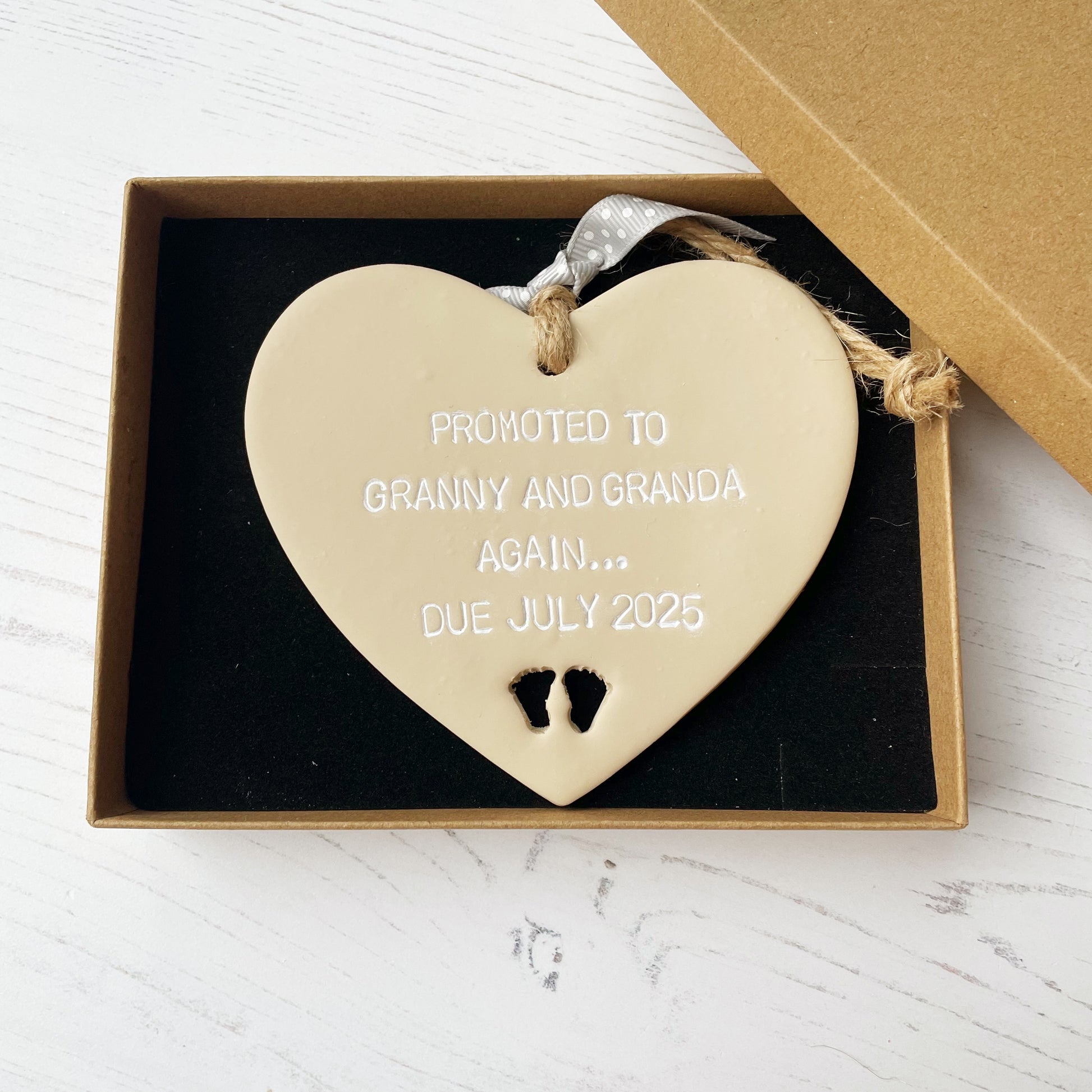 Beige clay hanging heart with baby feet cut out of the bottom and white personalisation, the heart is personalised with PROMOTED TO GRANNY AND GRANDA AGAIN… DUE JULY 2025