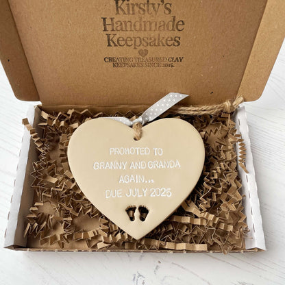 Beige clay hanging heart with baby feet cut out of the bottom and white personalisation, the heart is personalised with PROMOTED TO GRANNY AND GRANDA AGAIN… DUE JULY 2025