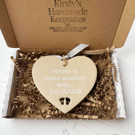 Beige clay hanging heart with baby feet cut out of the bottom and white personalisation, the heart is personalised with PROMOTED TO GRANNY AND GRANDA AGAIN… DUE JULY 2025