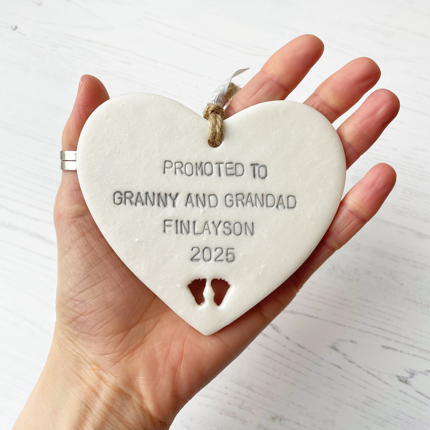 Pearlised white clay hanging heart with baby feet cut out of the bottom and grey personalisation, the heart is personalised with PROMOTED TO GRANNY AND GRANDAD FINLAYSON 2025