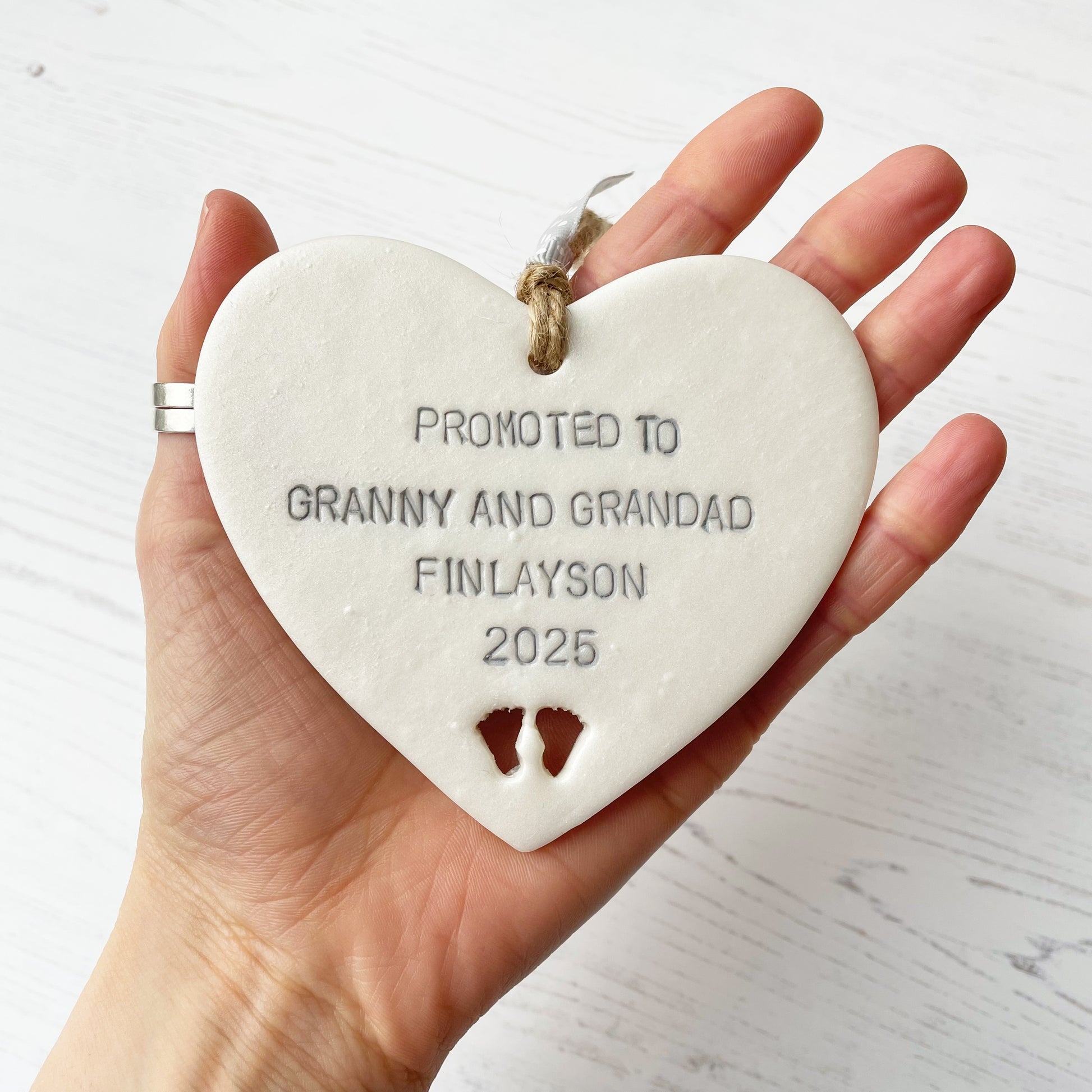 Pearlised white clay hanging heart with baby feet cut out of the bottom and grey personalisation, the heart is personalised with PROMOTED TO GRANNY AND GRANDAD FINLAYSON 2025