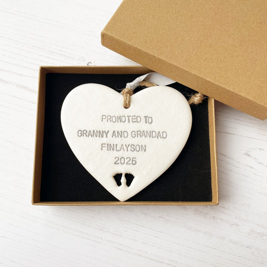 Pearlised white clay hanging heart with baby feet cut out of the bottom and grey personalisation, the heart is personalised with PROMOTED TO GRANNY AND GRANDAD FINLAYSON 2025