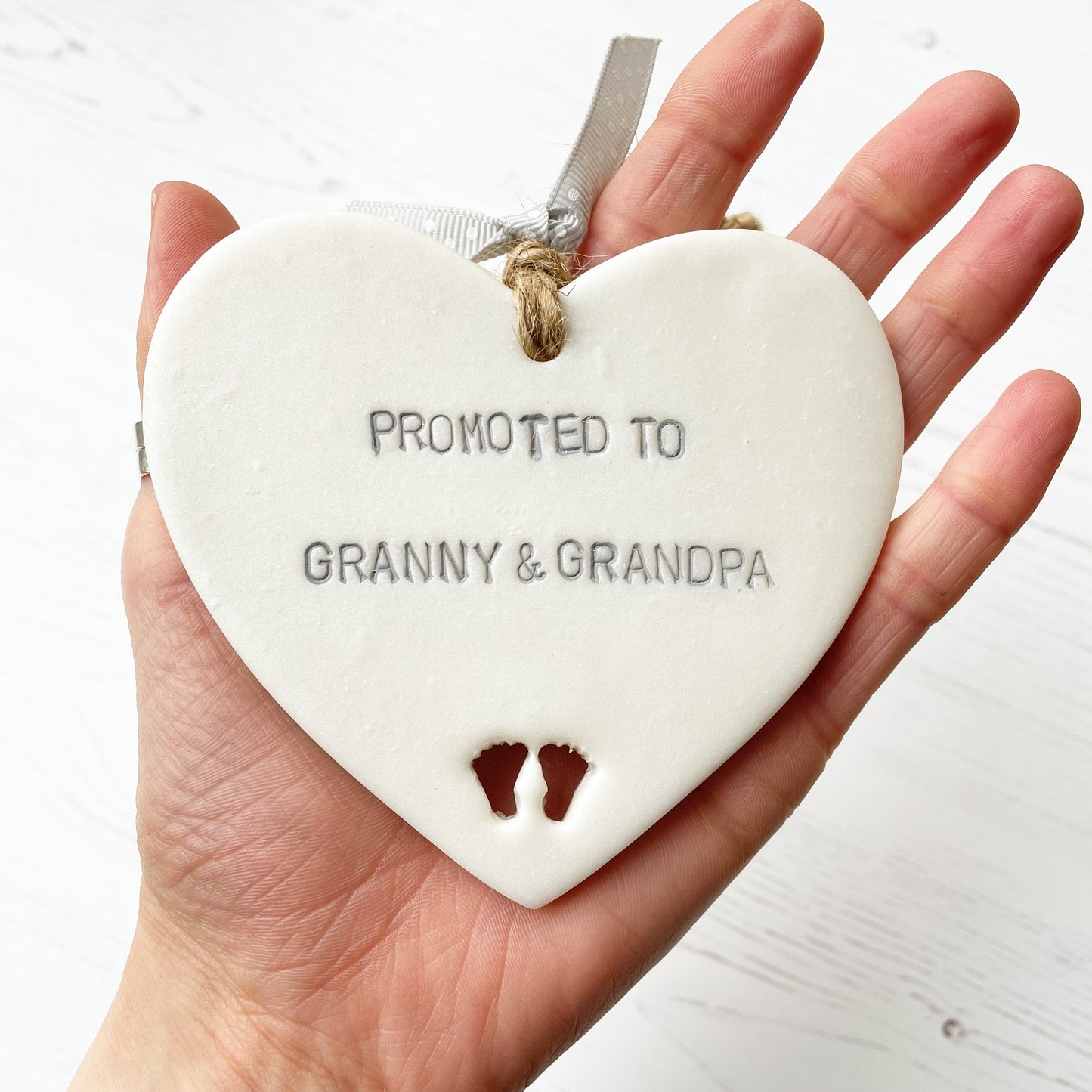 Pearlised white clay hanging heart with baby feet cut out of the bottom and grey personalisation, the heart is personalised with PROMOTED TO GRANNY & GRANDPA