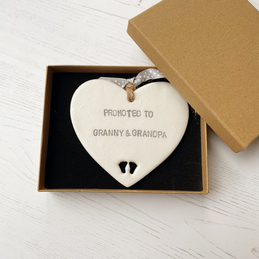 Pearlised white clay hanging heart with baby feet cut out of the bottom and grey personalisation, the heart is personalised with PROMOTED TO GRANNY & GRANDPA