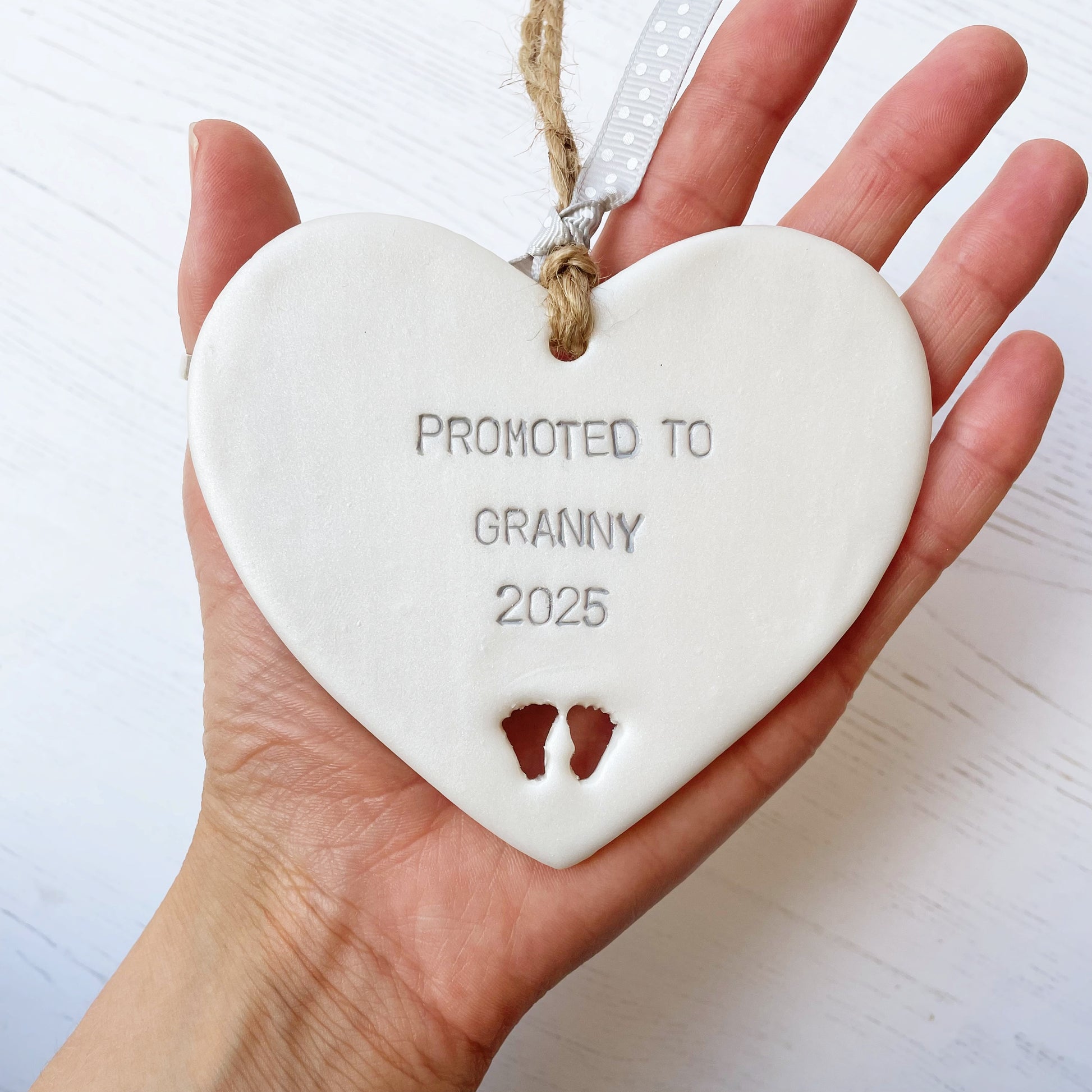 Pearlised white clay hanging heart with baby feet cut out of the bottom and grey personalisation, the heart is personalised with PROMOTED TO GRANNY 2025