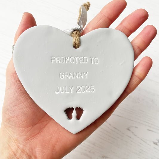 Grey clay hanging heart with baby feet cut out of the bottom and white personalisation, the heart is personalised with PROMOTED TO GRANNY 2025