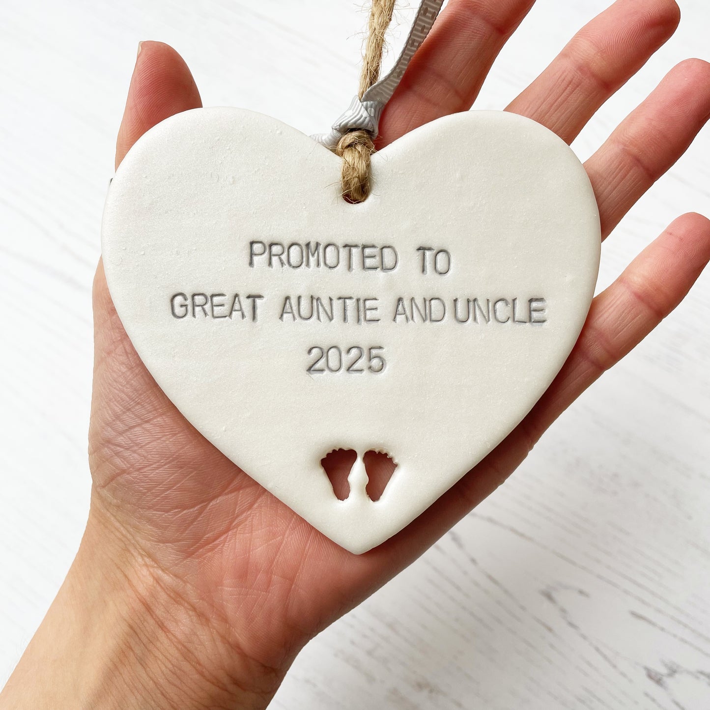 Pearlised white clay hanging heart with baby feet cut out of the bottom and grey personalisation, the heart is personalised with PROMOTED TO GREAT AUNTIE AND UNCLE 2025