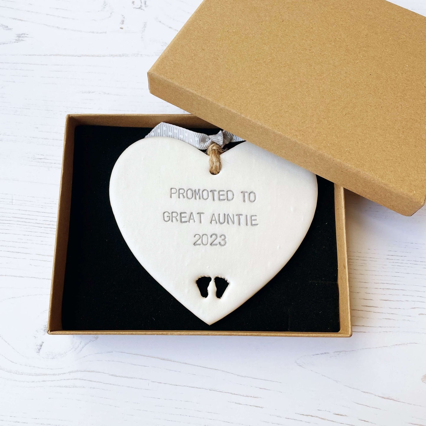 Pearlised white clay hanging heart with baby feet cut out of the bottom and grey personalisation, the heart is personalised with PROMOTED TO GREAT AUNTIE 2023