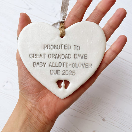 Pearlised white clay hanging heart with baby feet cut out of the bottom and grey personalisation, the heart is personalised with PROMOTED TO GREAT GRANDAD DAVE BABY ALLOTT-GLOVER DUE 2025