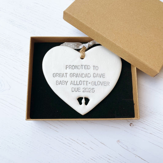 Pearlised white clay hanging heart with baby feet cut out of the bottom and grey personalisation, the heart is personalised with PROMOTED TO GREAT GRANDAD DAVE BABY ALLOTT-GLOVER DUE 2025