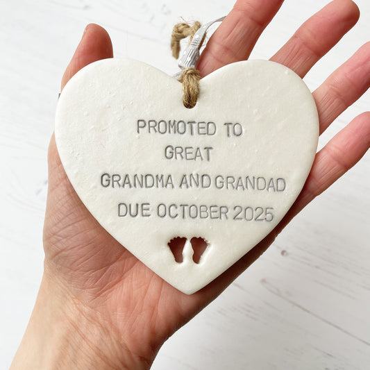 Pearlised white clay hanging heart with baby feet cut out of the bottom and grey personalisation, the heart is personalised with PROMOTED TO GREAT GRANDMA AND GRANDAD DUE OCTOBER 2025