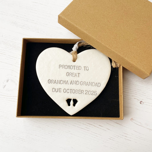 Pearlised white clay hanging heart with baby feet cut out of the bottom and grey personalisation, the heart is personalised with PROMOTED TO GREAT GRANDMA AND GRANDAD DUE OCTOBER 2025