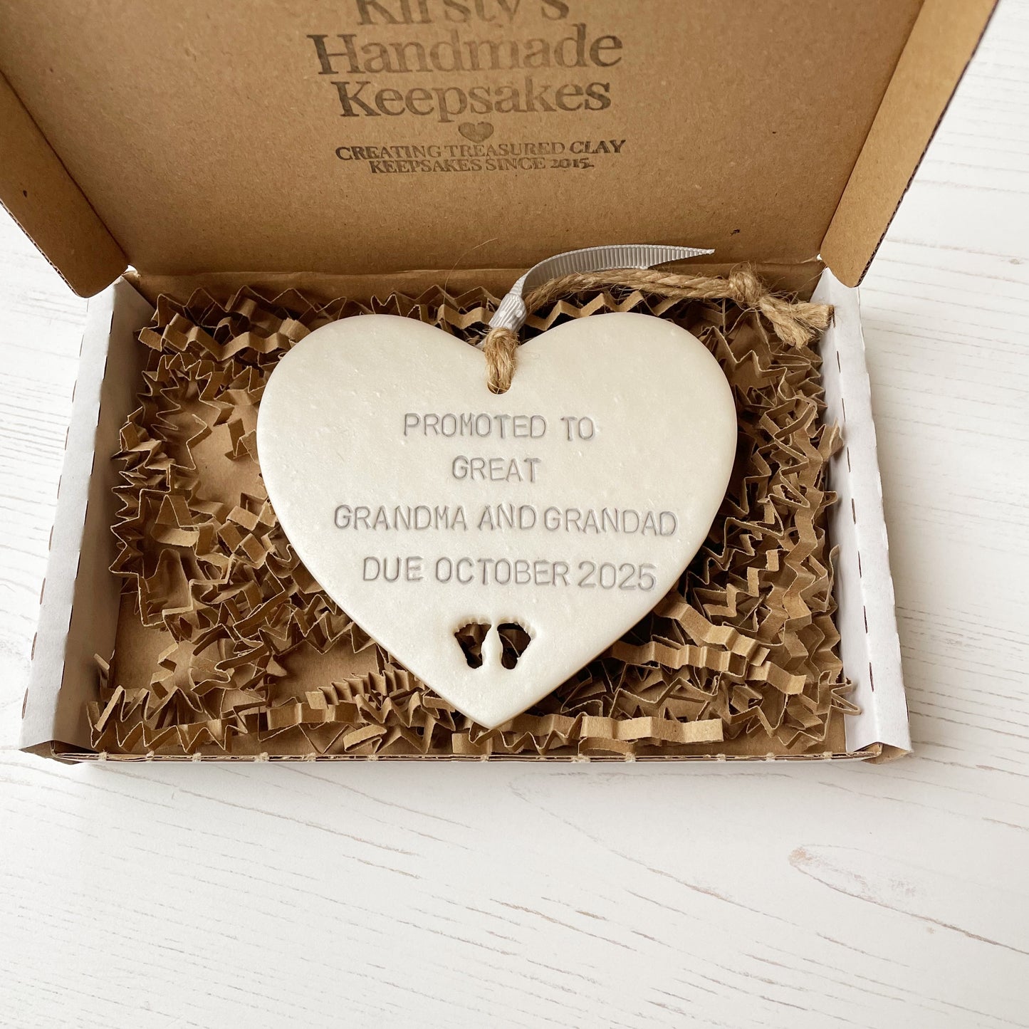 Pearlised white clay hanging heart with baby feet cut out of the bottom and grey personalisation, the heart is personalised with PROMOTED TO GREAT GRANDMA AND GRANDAD DUE OCTOBER 2025