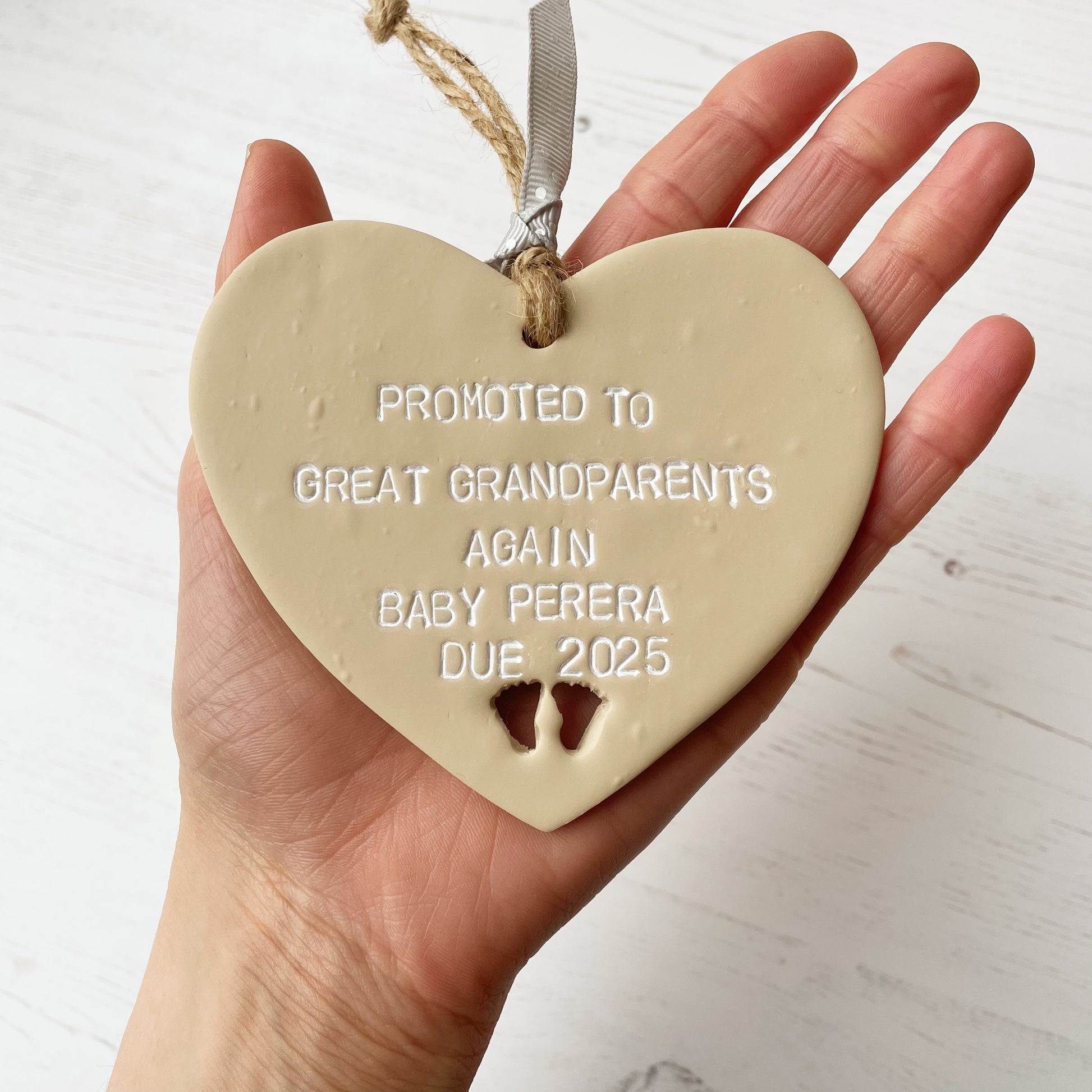 Beige clay hanging heart with baby feet cut out of the bottom and white personalisation, the heart is personalised with PROMOTED TO GREAT GRANDPARENTS AGAIN BABY PERERA DUE 2025