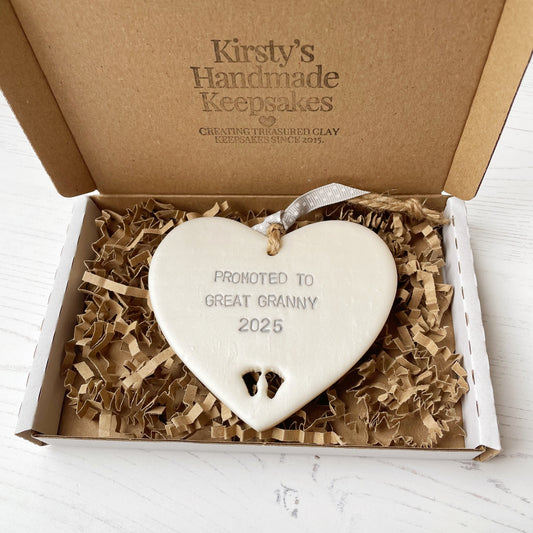 Pearlised white clay hanging heart with baby feet cut out of the bottom and grey personalisation, the heart is personalised with PROMOTED TO GREAT GRANNY 2025