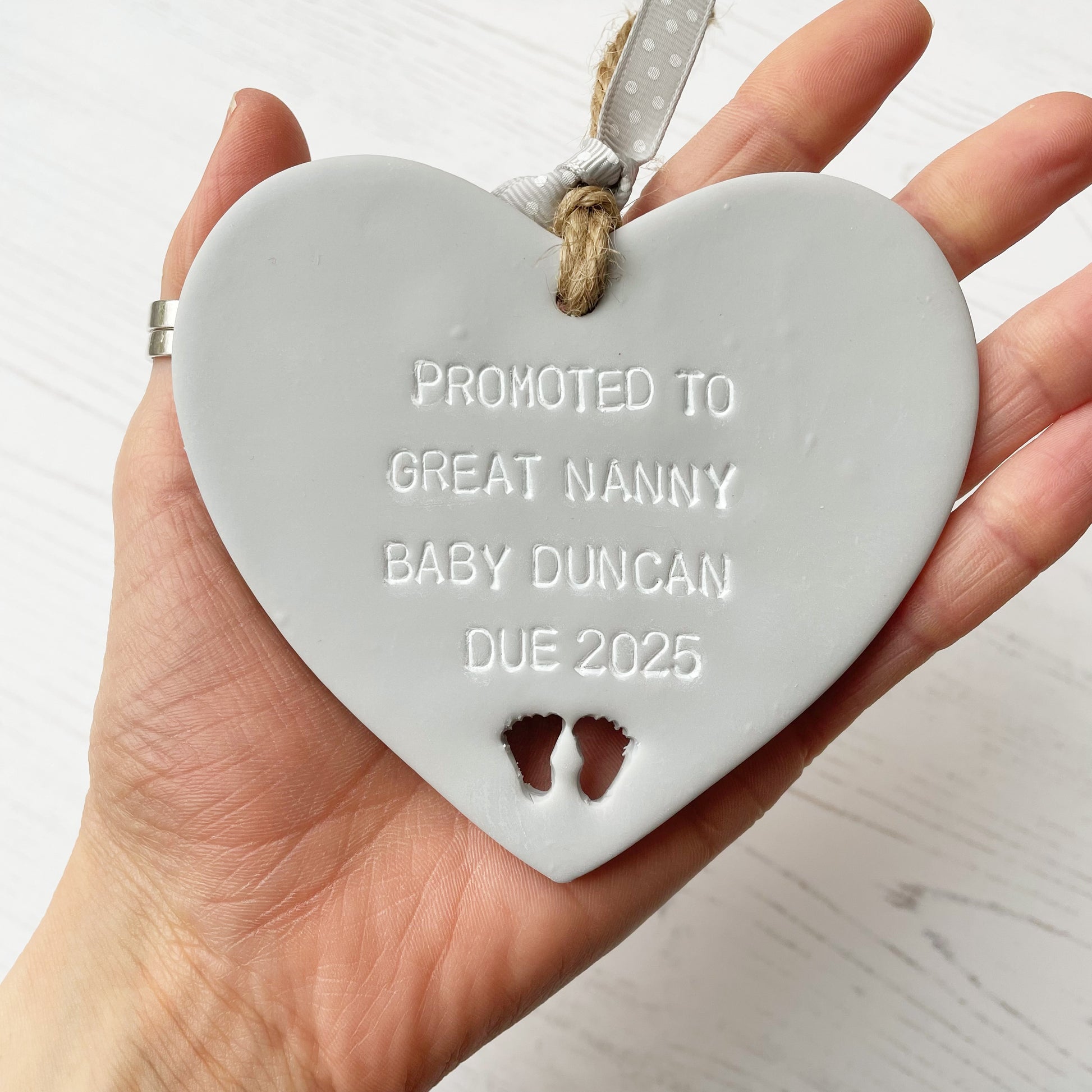 Grey clay hanging heart with baby feet cut out of the bottom and white personalisation, the heart is personalised with PROMOTED TO GREAT NANNY BABY DUNCAN DUE 2025