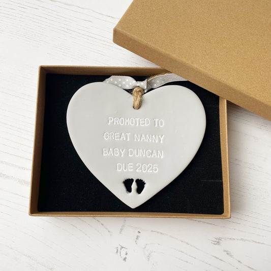 Grey clay hanging heart with baby feet cut out of the bottom and white personalisation, the heart is personalised with PROMOTED TO GREAT NANNY BABY DUNCAN DUE 2025