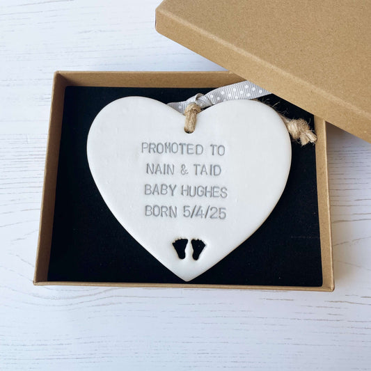 Pearlised white clay hanging heart with baby feet cut out of the bottom and grey personalisation, the heart is personalised with PROMOTED TO NAIN & TAID BABY HUGHES BORN 5/4/25