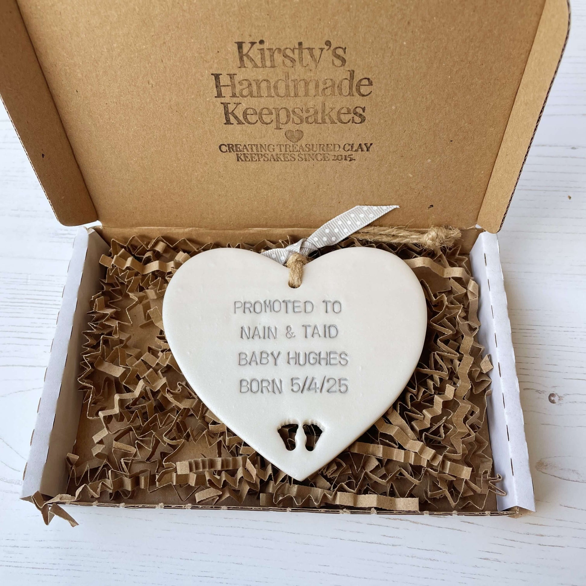 Pearlised white clay hanging heart with baby feet cut out of the bottom and grey personalisation, the heart is personalised with PROMOTED TO NAIN & TAID BABY HUGHES BORN 5/4/25