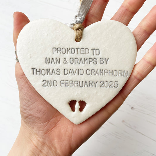 Pearlised white clay hanging heart with baby feet cut out of the bottom and grey personalisation, the heart is personalised with PROMOTED TO NAN & GRAMPS BY THOMAS DAVID CRAMPHORN 2ND FEBRUARY 2025