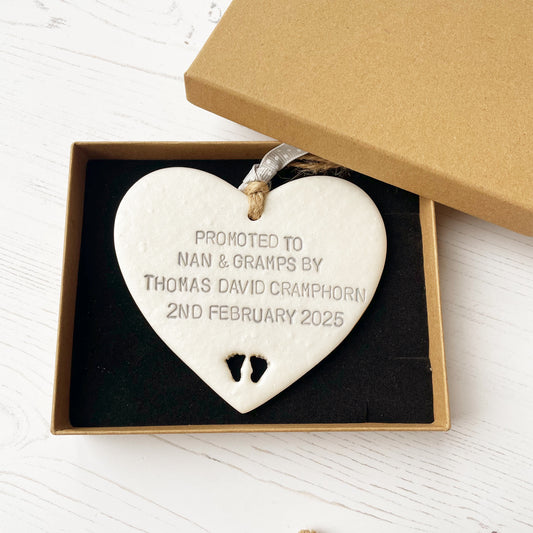 Pearlised white clay hanging heart with baby feet cut out of the bottom and grey personalisation, the heart is personalised with PROMOTED TO NAN & GRAMPS BY THOMAS DAVID CRAMPHORN 2ND FEBRUARY 2025