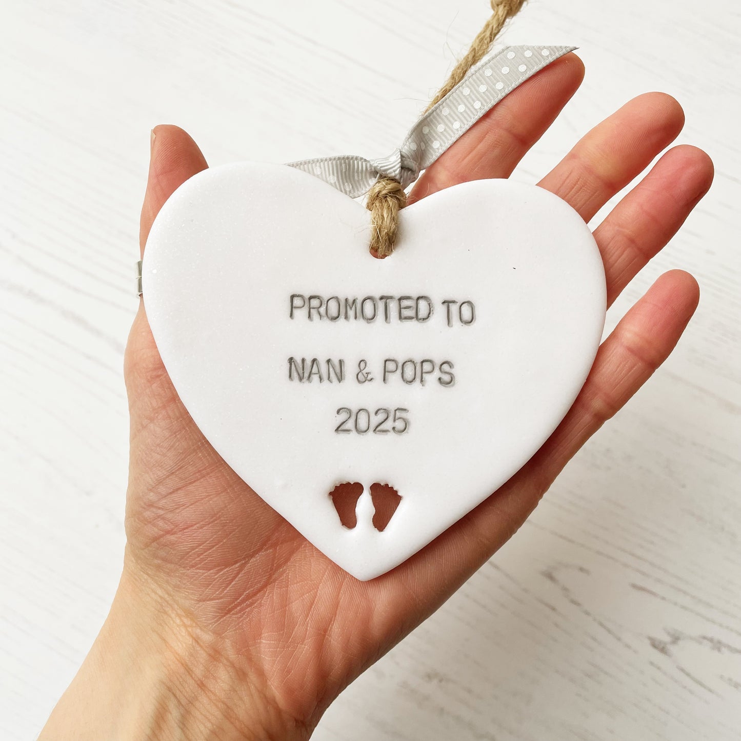 Pearlised white clay hanging heart with baby feet cut out of the bottom and grey personalisation, the heart is personalised with PROMOTED TO NAN AND POPS 2025