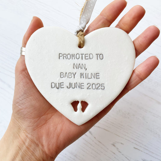 Pearlised white clay hanging heart with baby feet cut out of the bottom and grey personalisation, the heart is personalised with PROMOTED TO NAN, BABY MILNE DUE JUNE 2025