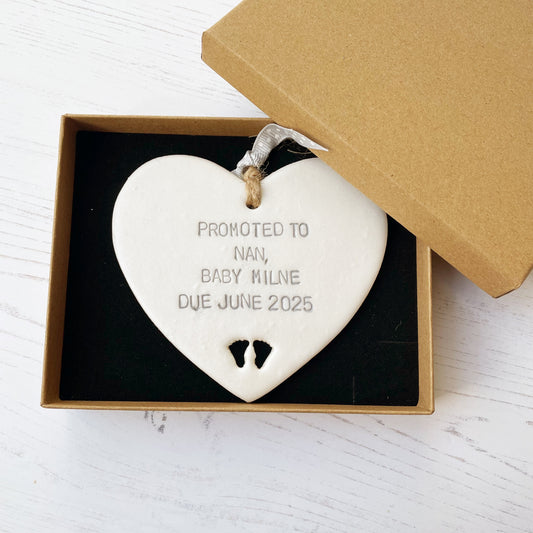 Pearlised white clay hanging heart with baby feet cut out of the bottom and grey personalisation, the heart is personalised with PROMOTED TO NAN, BABY MILNE DUE JUNE 2025