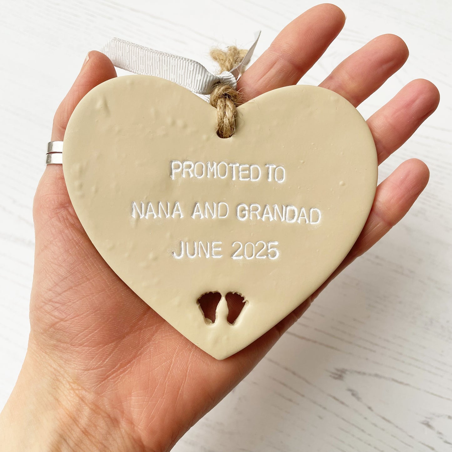 Beige clay hanging heart with baby feet cut out of the bottom and white personalisation, the heart is personalised with PROMOTED TO NANA AND GRANDAD JUNE 2025