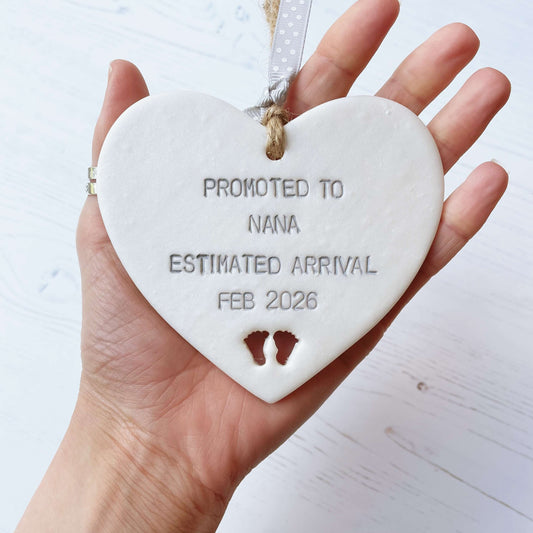 Pearlised white clay hanging heart with baby feet cut out of the bottom and grey personalisation, the heart is personalised with PROMOTED TO NANA ESTIMATED ARRIVAL FEB 2026