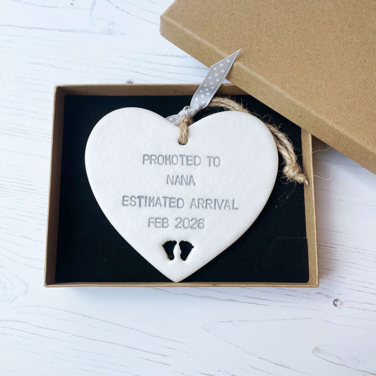 Pearlised white clay hanging heart with baby feet cut out of the bottom and grey personalisation, the heart is personalised with PROMOTED TO NANA ESTIMATED ARRIVAL FEB 2026