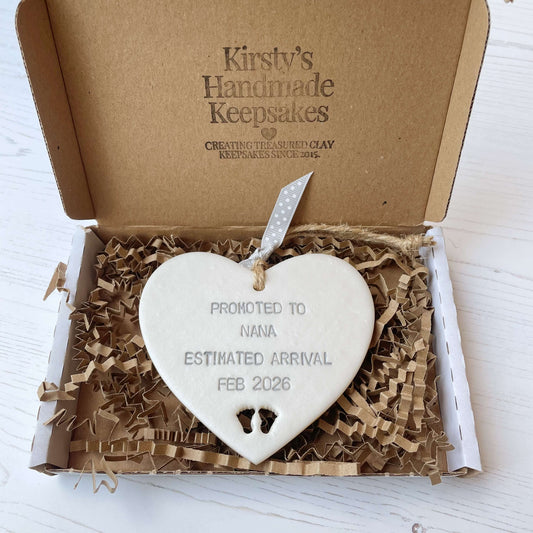 Pearlised white clay hanging heart with baby feet cut out of the bottom and grey personalisation, the heart is personalised with PROMOTED TO NANA ESTIMATED ARRIVAL FEB 2026