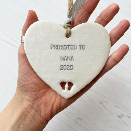 Pearlised white clay hanging heart with baby feet cut out of the bottom and grey personalisation, the heart is personalised with PROMOTED TO NANA 2025