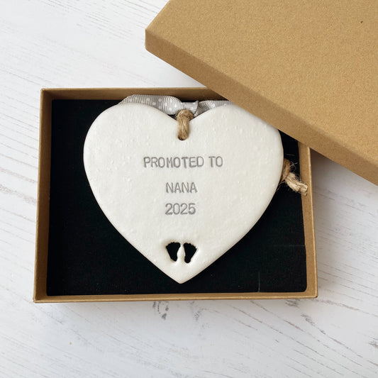 Pearlised white clay hanging heart with baby feet cut out of the bottom and grey personalisation, the heart is personalised with PROMOTED TO NANA 2025