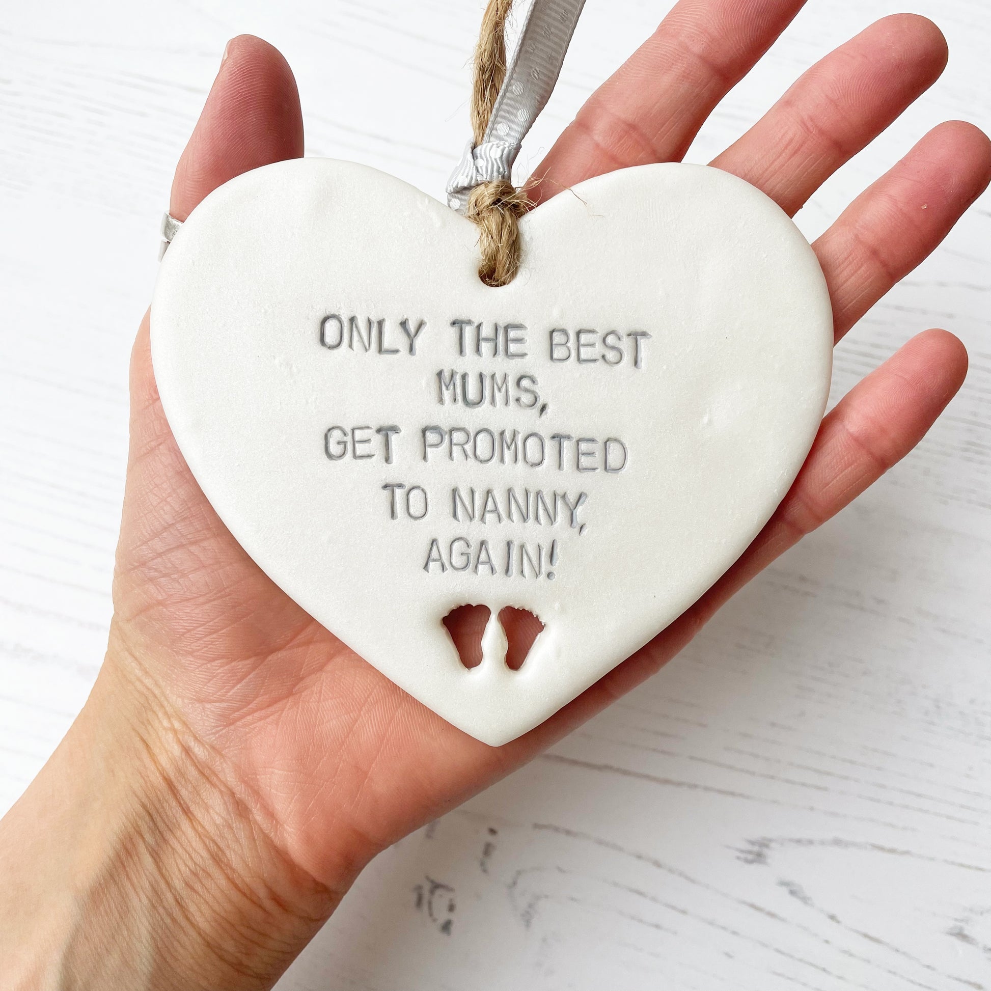 Pearlised white clay hanging heart with baby feet cut out of the bottom and grey personalisation, the heart is personalised with ONLY THE BEST MUMS, GET PROMOTED TO NANNY, AGAIN!