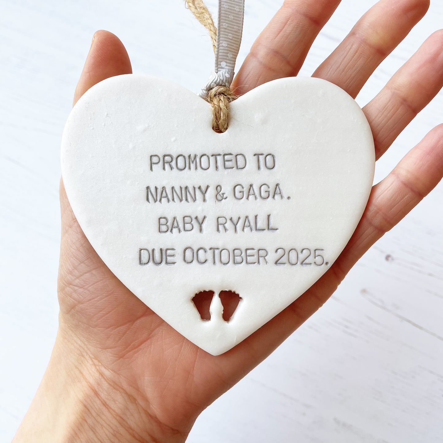 Pearlised white clay hanging heart with baby feet cut out of the bottom and grey personalisation, the heart is personalised with PROMOTED TO NANNY & GAGA. BABY RYALL DUE OCTOBER 2025.