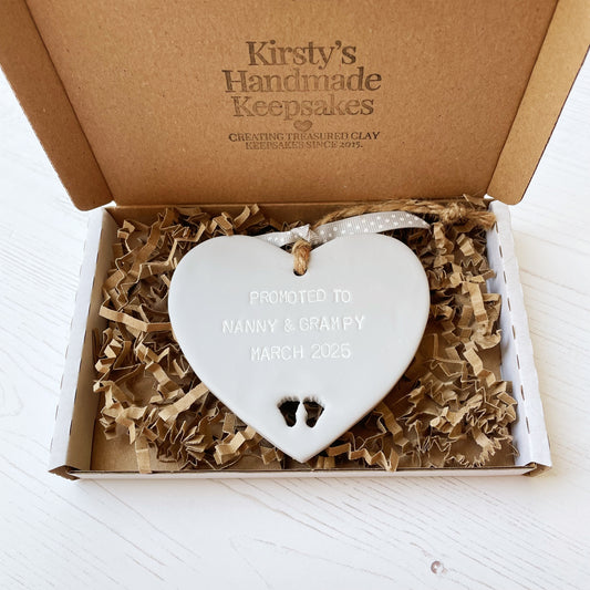 Promoted to Grandparents Grey clay hanging heart with baby feet cut out of the bottom and white personalisation, the heart is personalised with PROMOTED TO NANNY & GRAMPY MARCH 2025