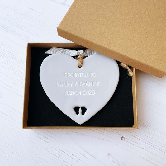 Promoted to Grandparents Grey clay hanging heart with baby feet cut out of the bottom and white personalisation, the heart is personalised with PROMOTED TO NANNY & GRAMPY MARCH 2025
