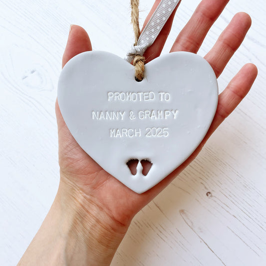 Promoted to Grandparents Grey clay hanging heart with baby feet cut out of the bottom and white personalisation, the heart is personalised with PROMOTED TO NANNY & GRAMPY MARCH 2025