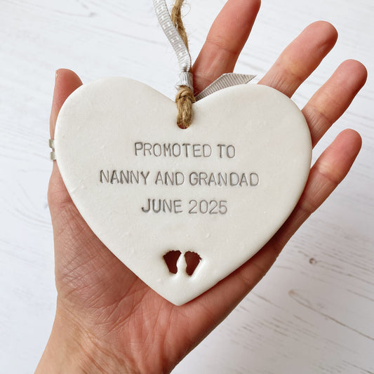 Pearlised white clay hanging heart with baby feet cut out of the bottom and grey personalisation, the heart is personalised with PROMOTED TO NANNY AND GRANDAD JUNE 2025