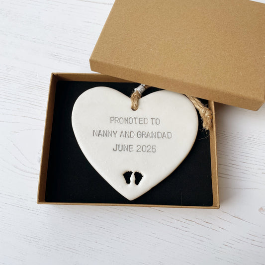 Pearlised white clay hanging heart with baby feet cut out of the bottom and grey personalisation, the heart is personalised with PROMOTED TO NANNY AND GRANDAD JUNE 2025