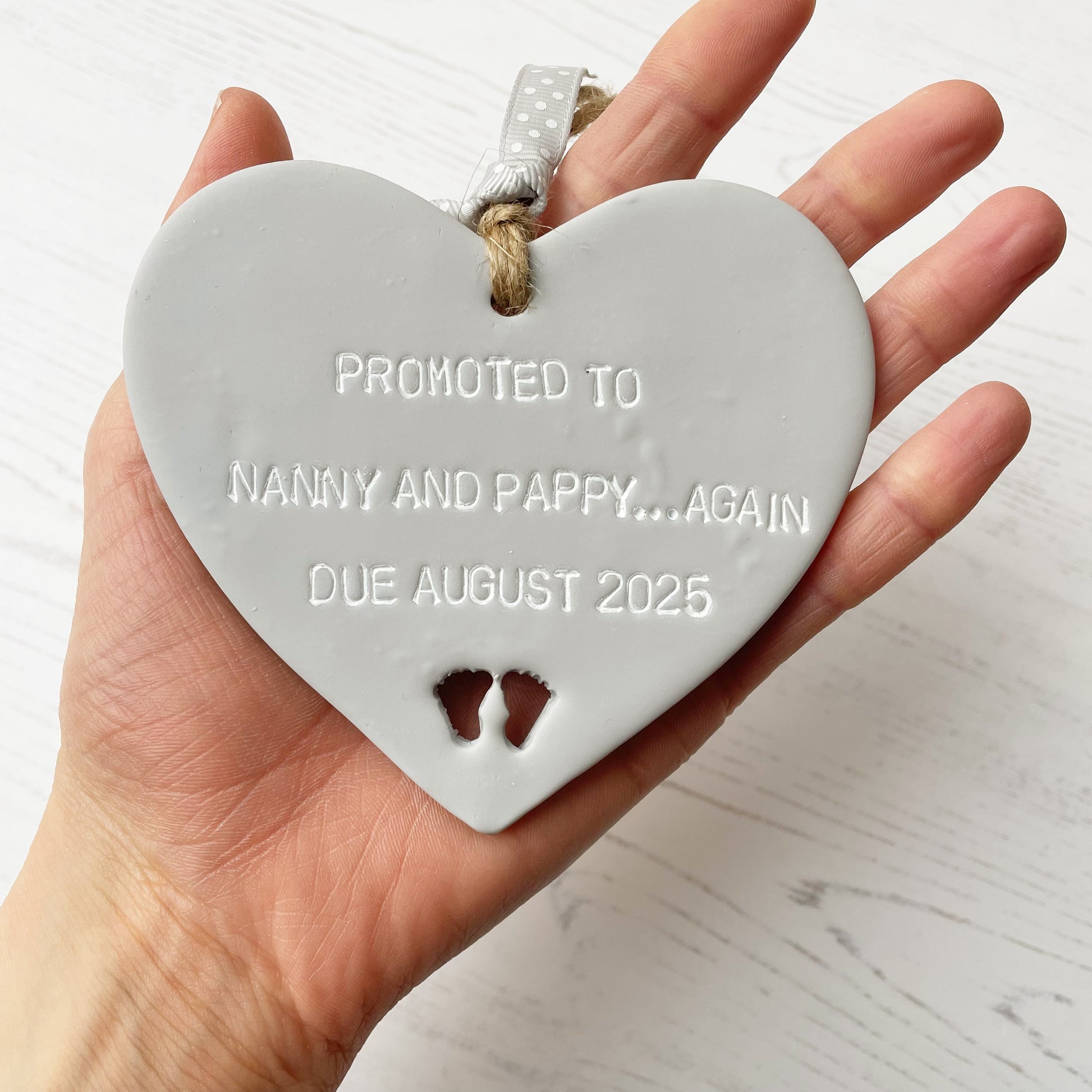 Grey clay hanging heart with baby feet cut out of the bottom and grey personalisation, the heart is personalised with PROMOTED TO NANNY AND PAPPY… AGAIN DUE AUGUST 2025